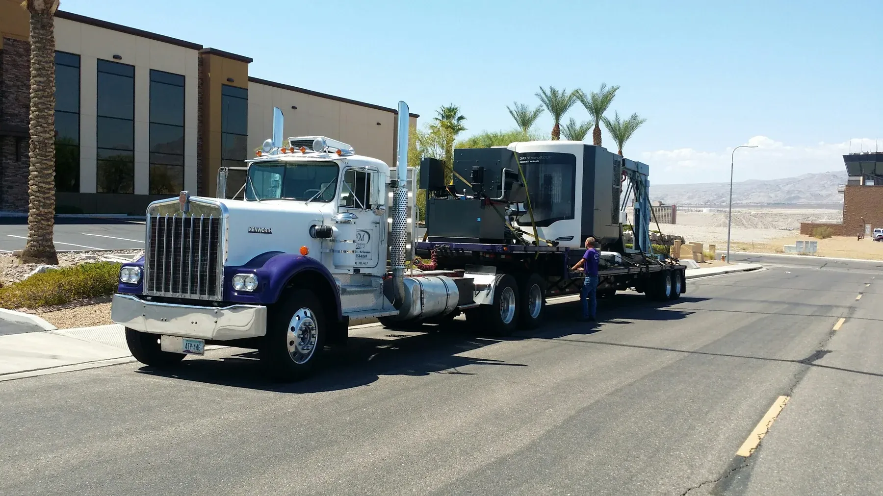A C&M Rigging Kenworth hauling a DMG Mori DMU 85 monoBLOCK CNC machining center strapped to a flatbed trailer, with a crew member in a purple shirt standing alongside.