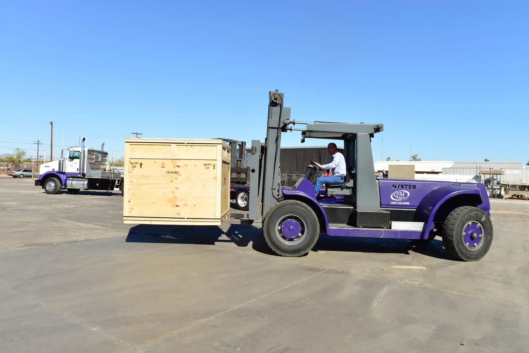 A C&M Rigging operator driving a purple Hyster forklift carrying a large plywood shipping crate across the Phoenix yard.